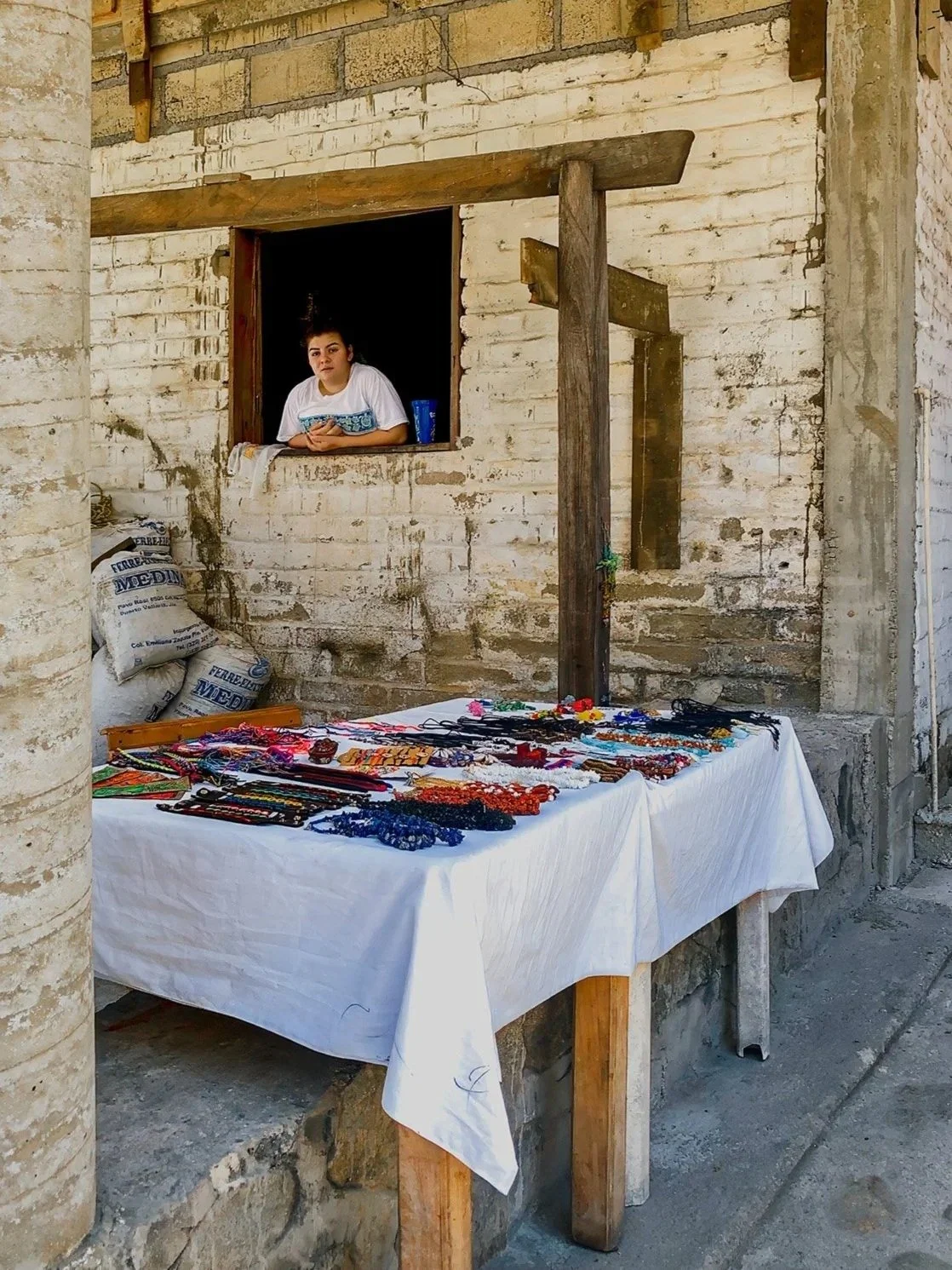 A young woman watching from a window above a table covered with colorful jewelry at an outdoor market stall.