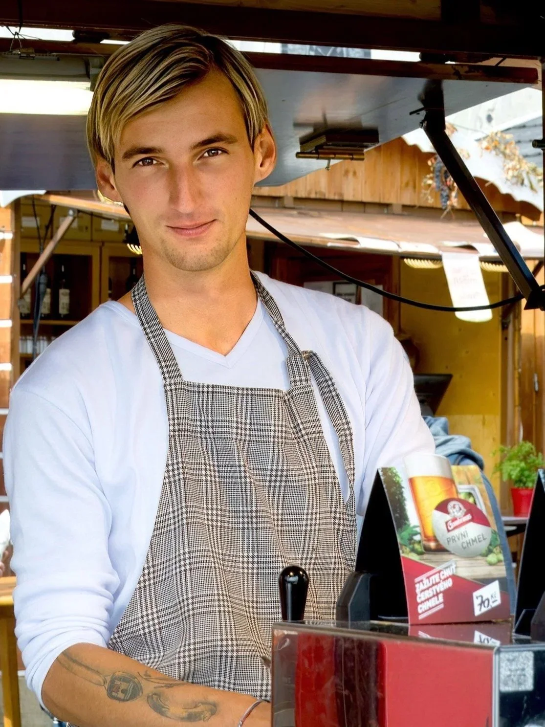 A young man with blond hair, wearing a white shirt and a plaid apron, standing behind a counter at an outdoor food stand.