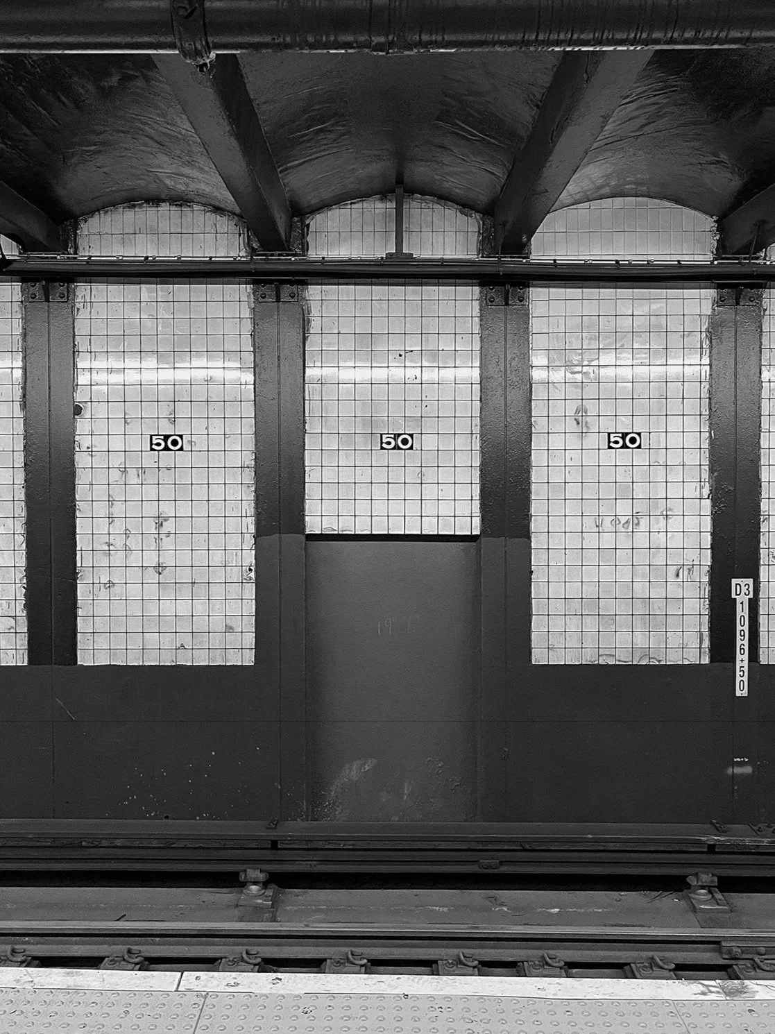  subway or station platform, showing the edge of the platform with metal tracks in the foreground, and a wall with three large, grid-like panels marked with '50' in black on white backgrounds.