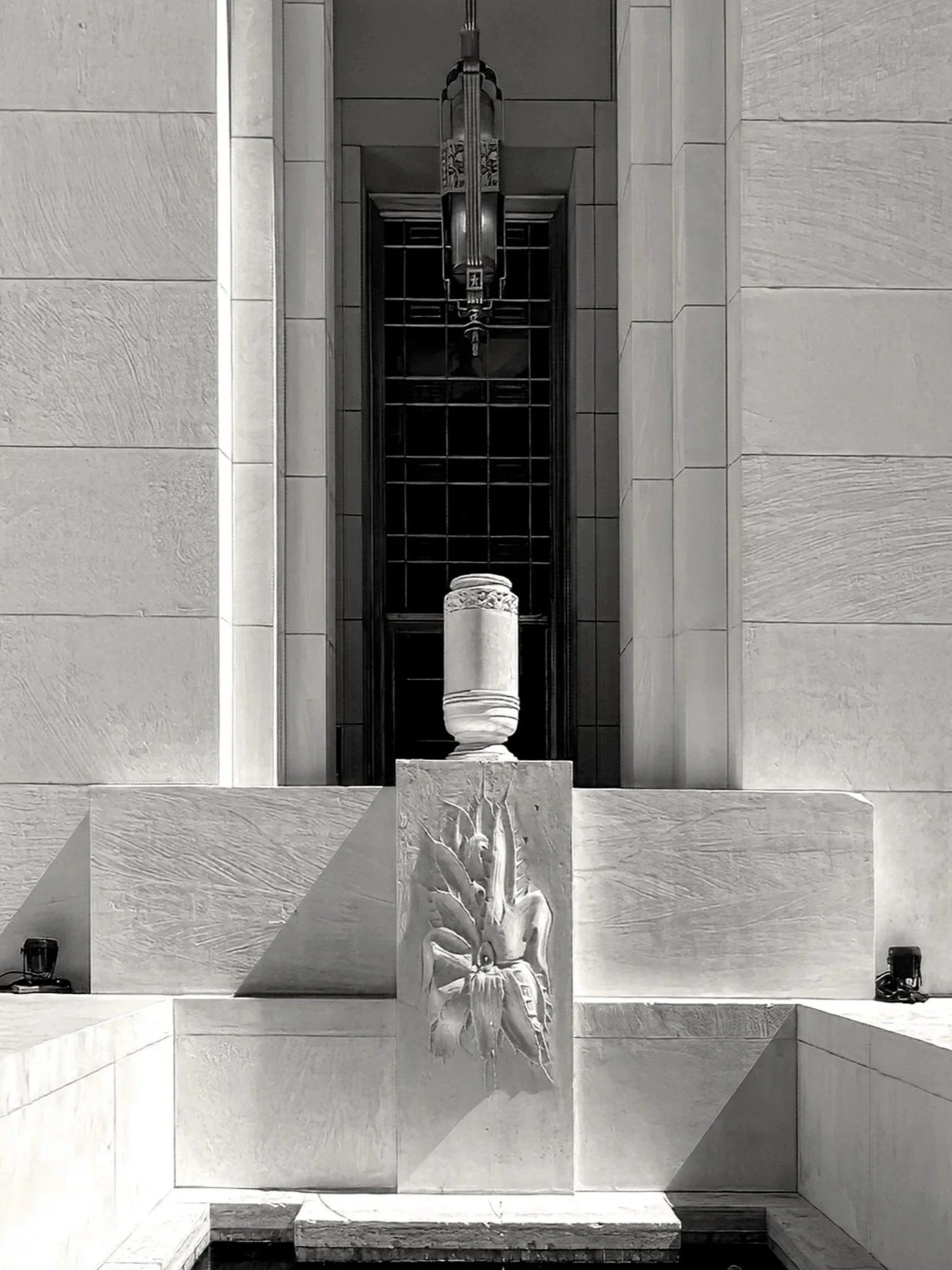 A black-and-white photo of a monument with a classical style. It features a tall cylindrical urn on a pedestal with carved floral designs.