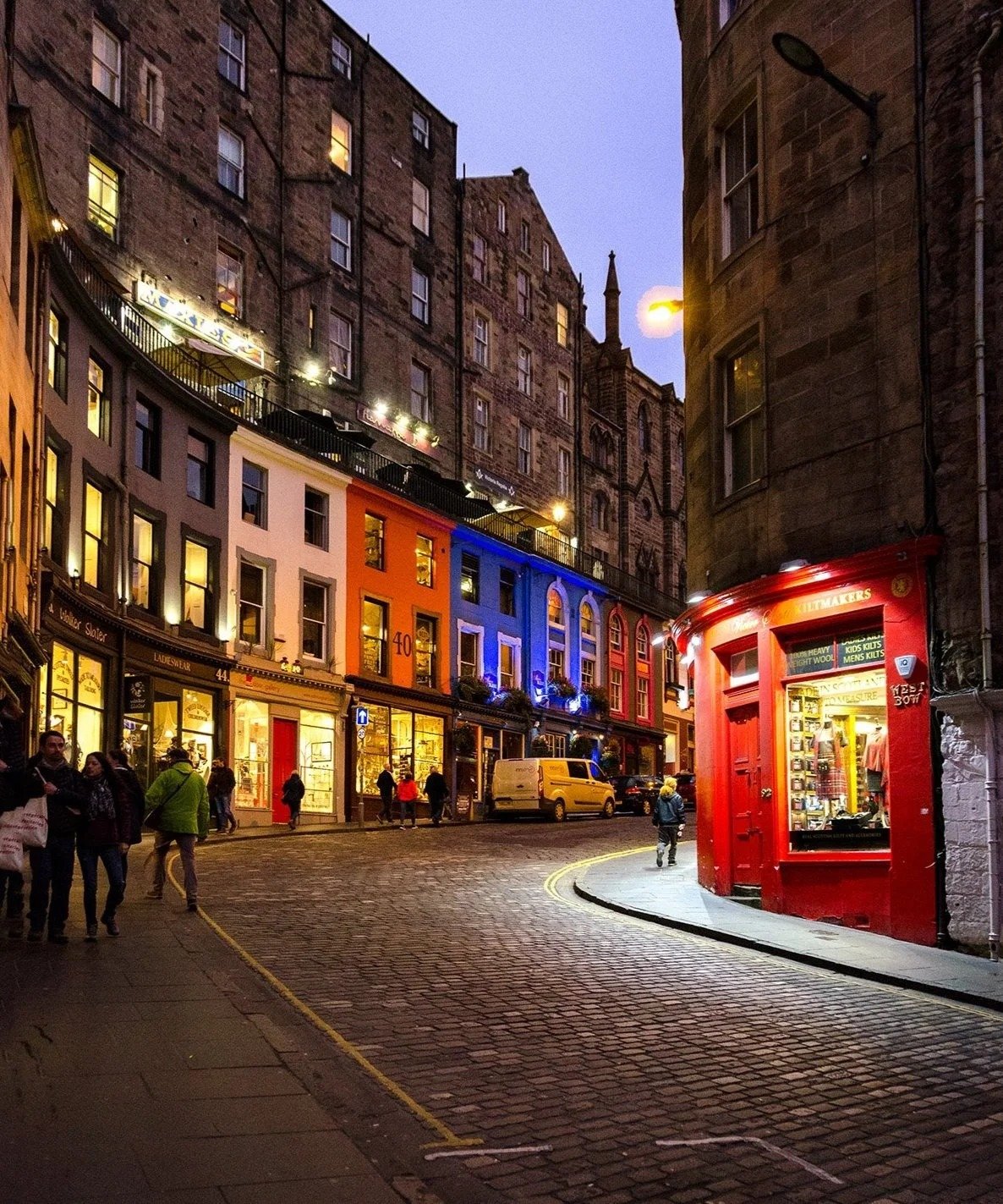Nighttime view of a cobblestone street with colorful buildings, people walking, and a red storefront on the corner.