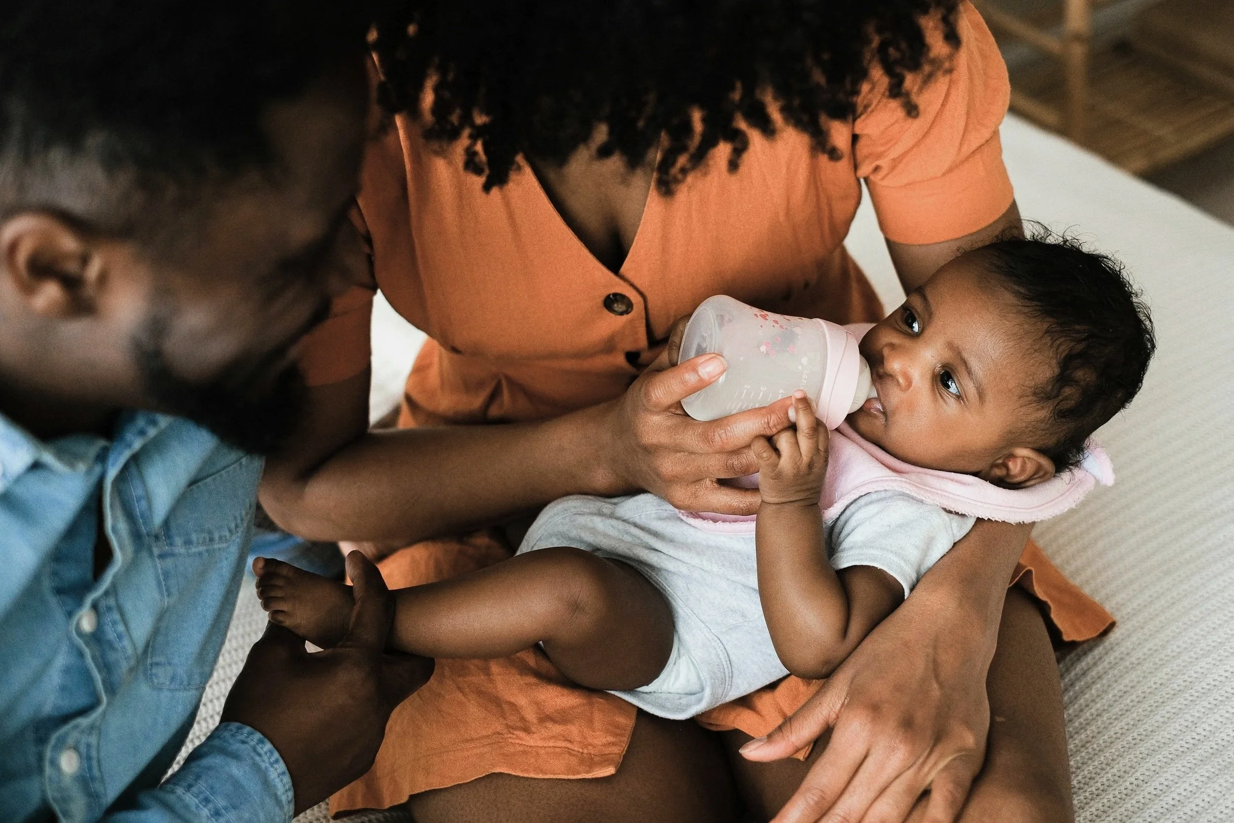 A woman feeds a baby with a bottle while sitting on a bed, with a man beside her helping to hold the baby.