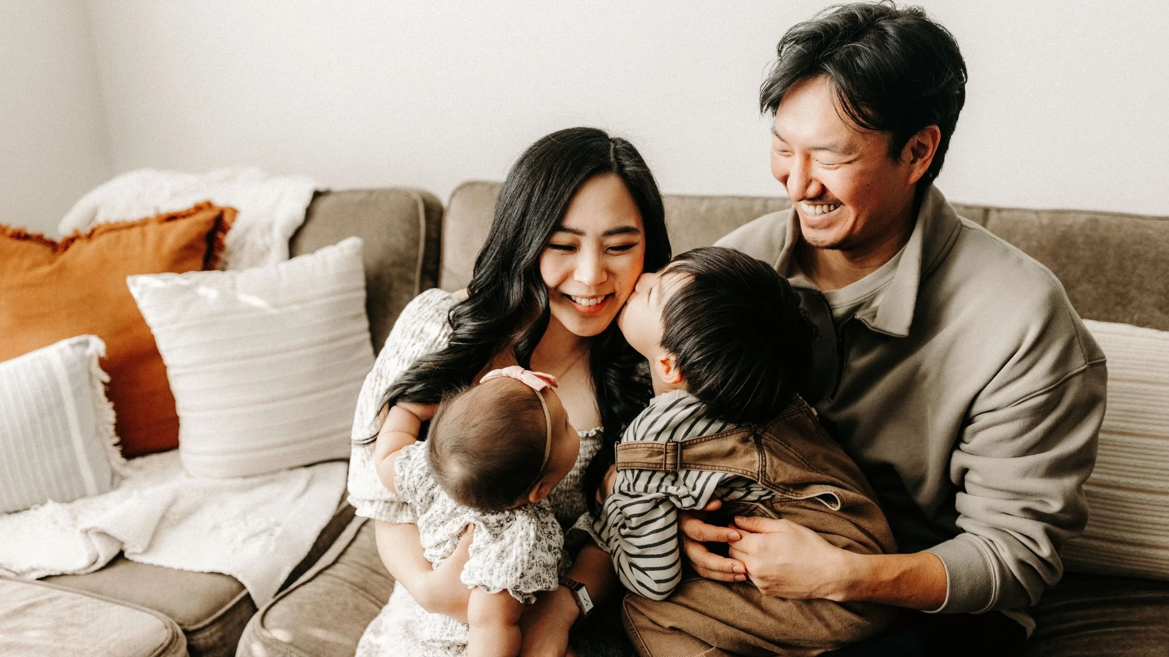 A happy family of four sitting on a beige couch in a living room, smiling and sharing affectionate moments with their two young children.