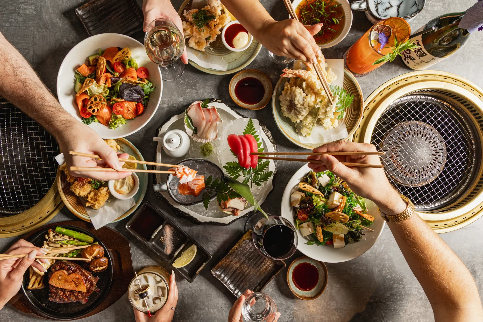 A top-down view of a Japanese-style barbecue feast featuring various dishes including sashimi, tempura, salad, grilled meat, and vegetables, with drinks and dipping sauces on a table.