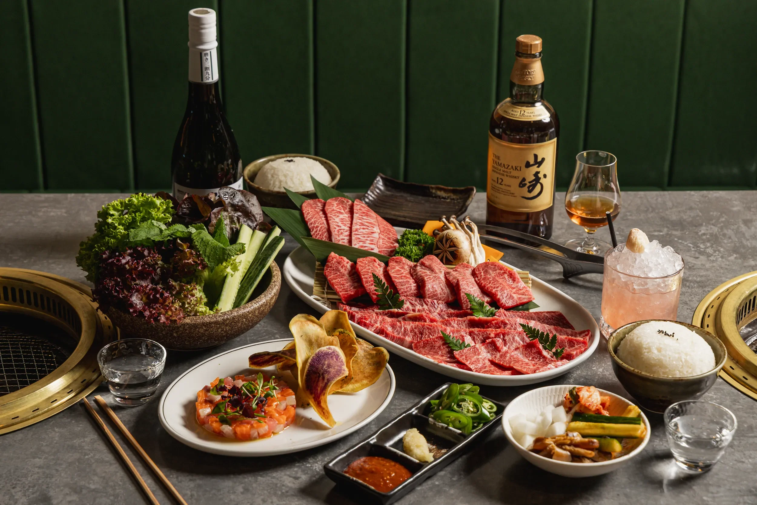 A table set for a Japanese barbecue meal with various dishes including marbled beef, vegetables, rice, and sauces, along with drinks including whiskey, wine, and a cocktail. The table has a built-in grill and green wall background.