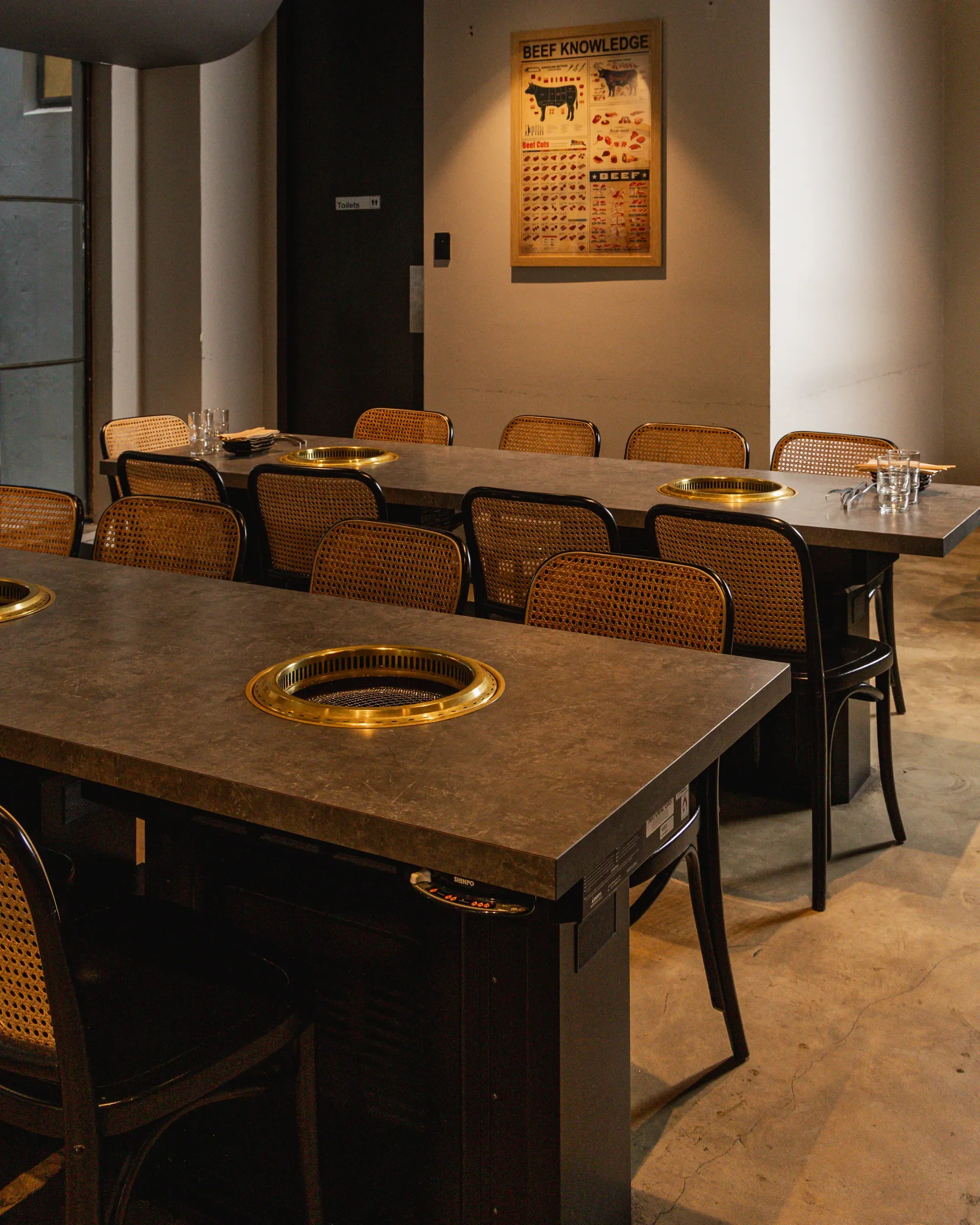 Empty restaurant with dark tables, chairs with woven backs, and built-in grill openings in the tables, set with glasses and utensils. There is a 'Toilets' sign on the door in the background and a 'Beef Knowledge' poster on the wall.