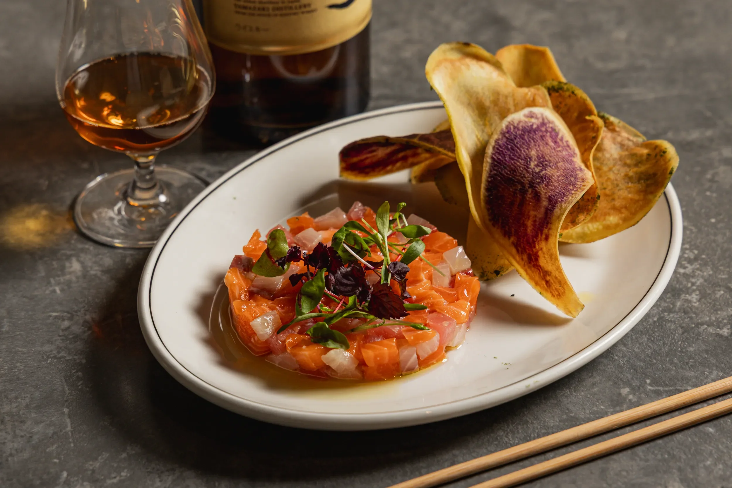 Plate with tartare topped with microgreens and served with crispy chips; glass of rosé wine and a bottle in the background.