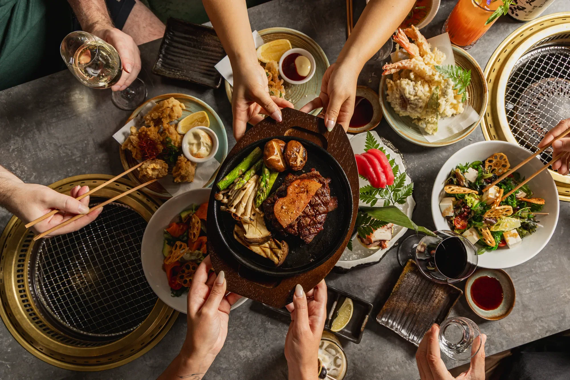 Table with various dishes including a sizzling platter of steak with vegetables, fried chicken with dipping sauces, a fresh salad, and beverages, with people reaching in to share the food in a Japanese-style barbecue restaurant.