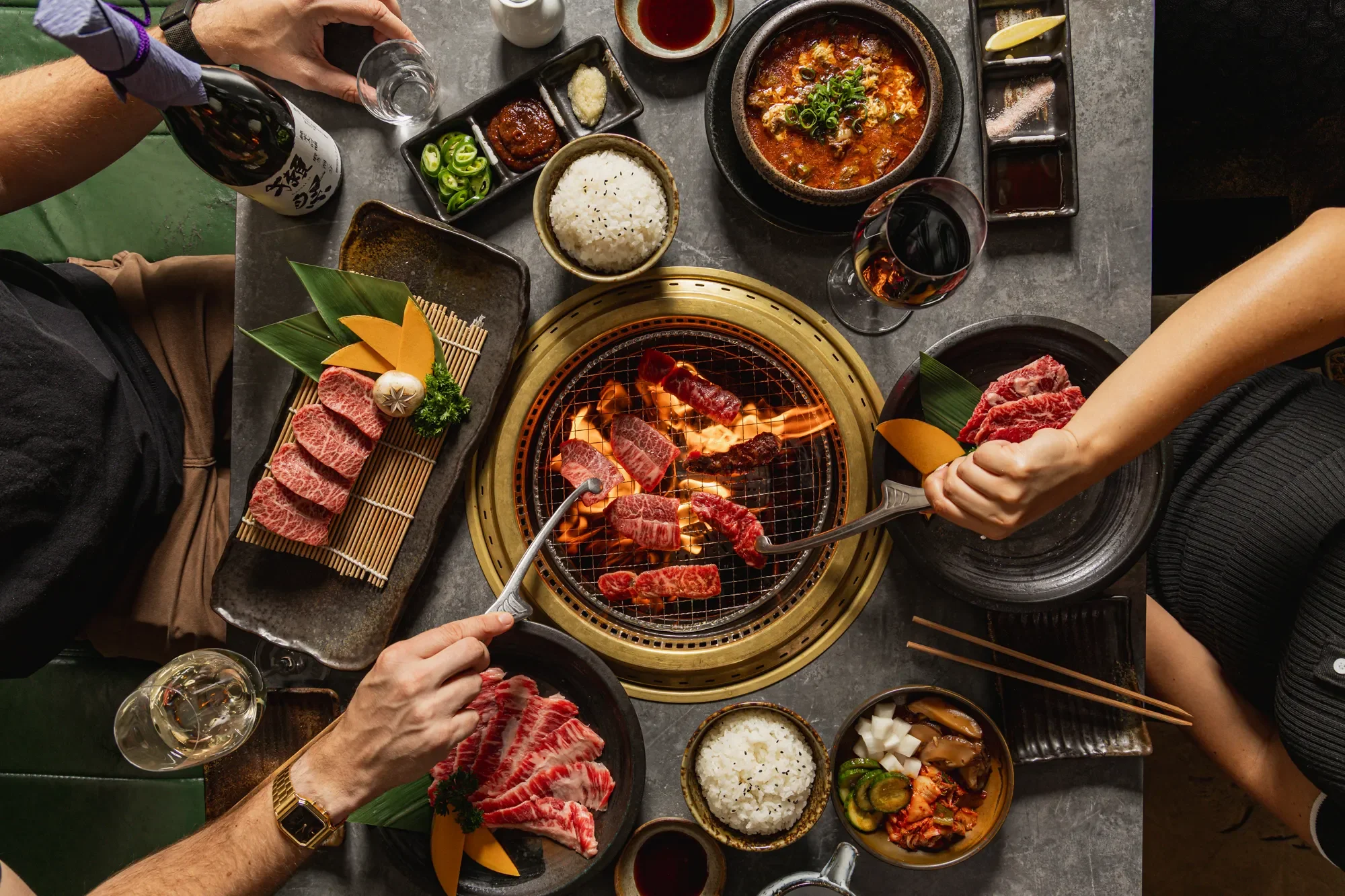 People cooking and eating Korean barbecue at a table, with raw meat being grilled over an open flame, surrounded by side dishes, bowls, glasses of drinks, and chopsticks.