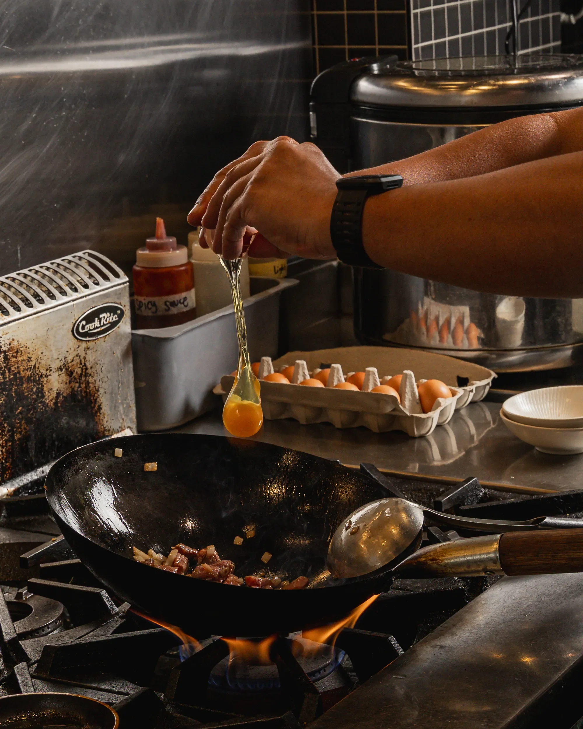 A person cracking eggs into a black frying pan over a gas stove, with other kitchen items visible in the background.