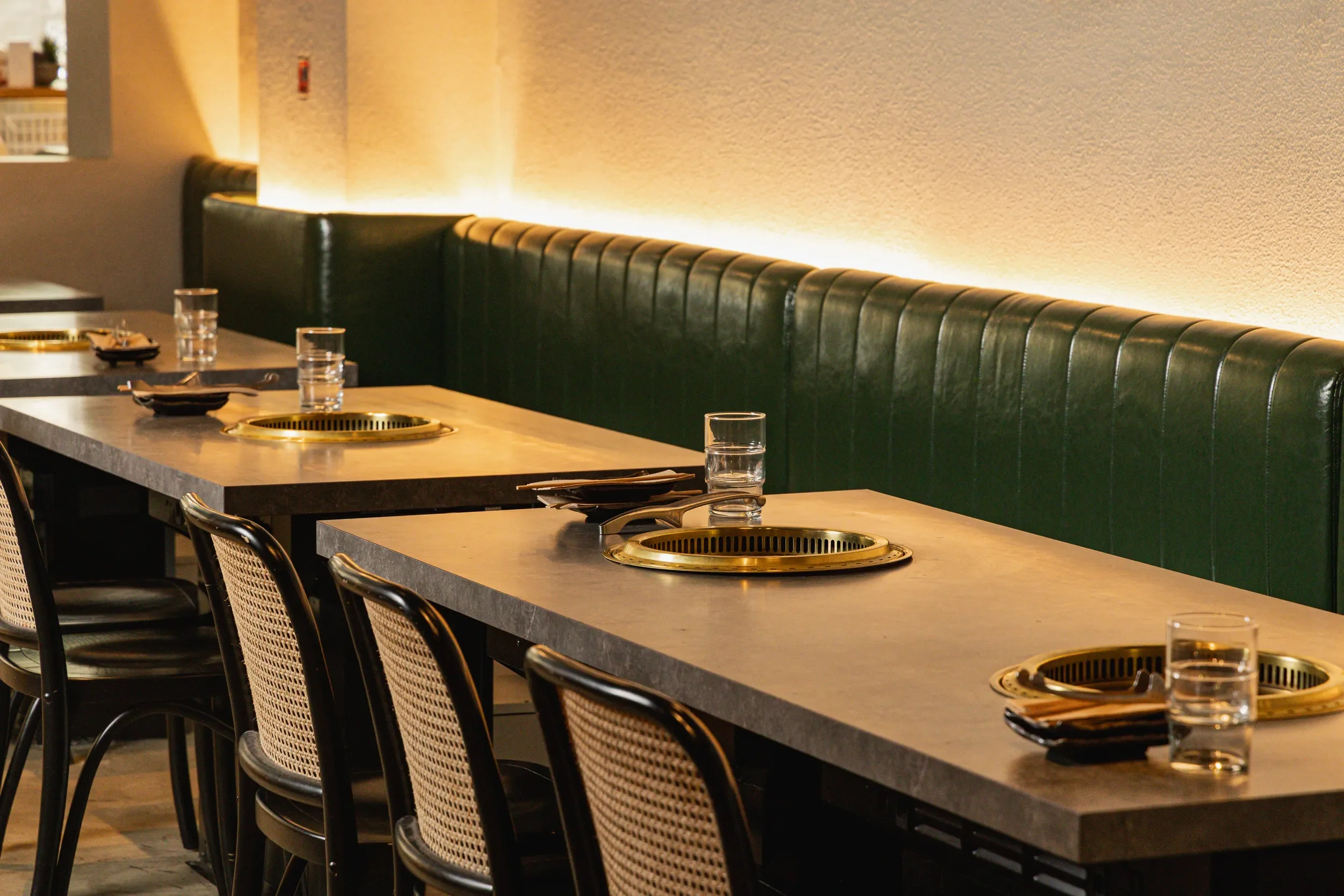 Empty restaurant table with built-in grill in a modern dining area, featuring a long green leather booth, chairs, glasses of water, utensils, and napkins.