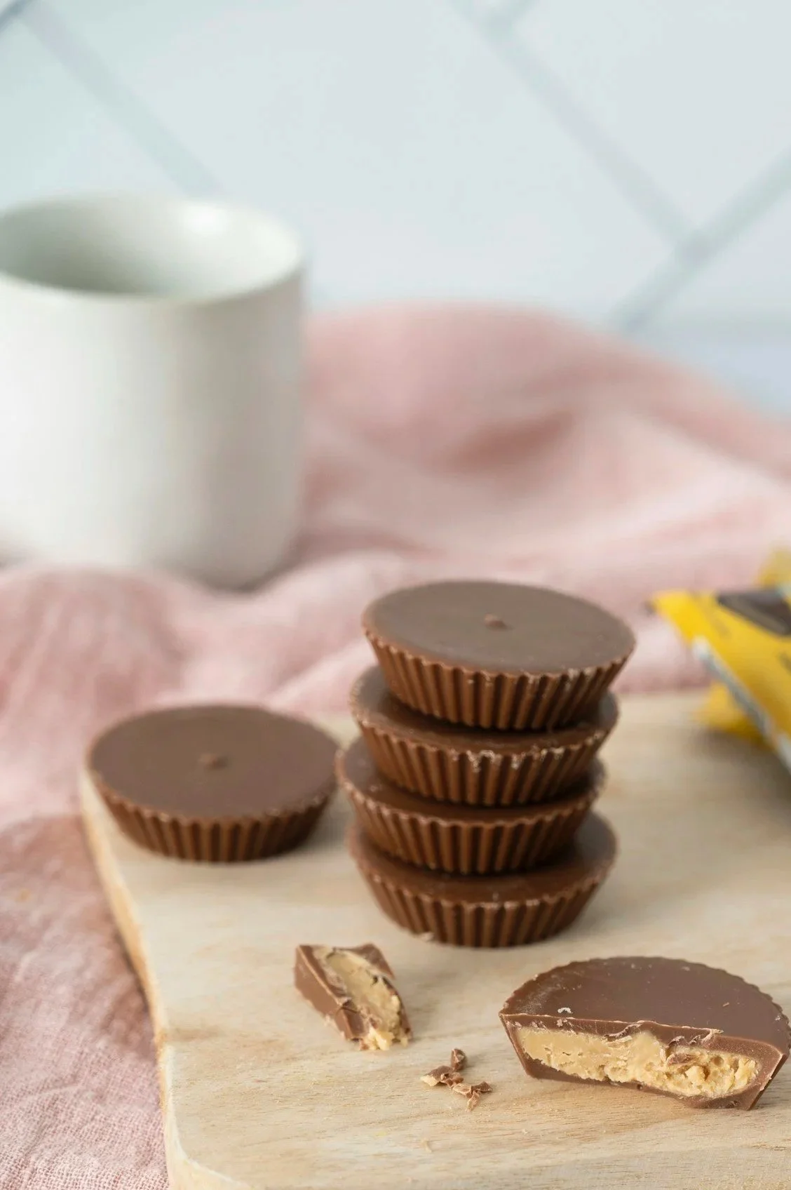 A stack of four chocolates with peanut filling, one broken piece showing filling inside, on a light wooden board. A white mug with a dark beverage is in the background, with a pink cloth underneath and a yellow chocolate wrapper nearby.