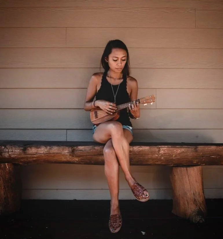 A young woman sitting on a rustic wooden bench, playing a ukulele, in front of a wooden paneled wall, wearing a black tank top and shorts.