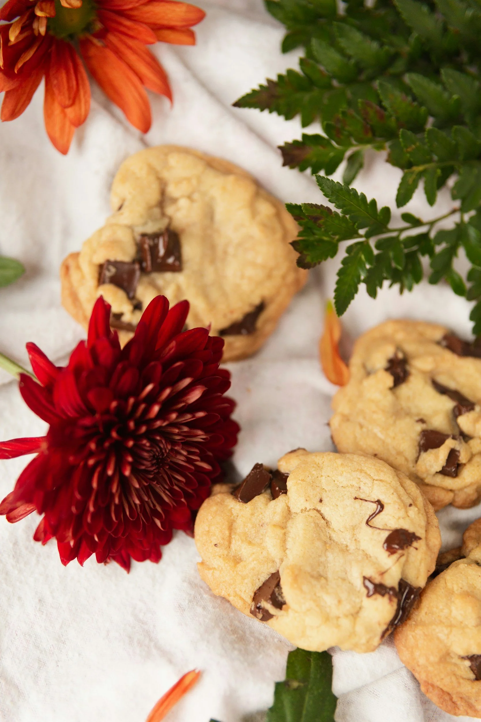Chocolate chip cookies with red and orange flowers and green leaves on a white surface.