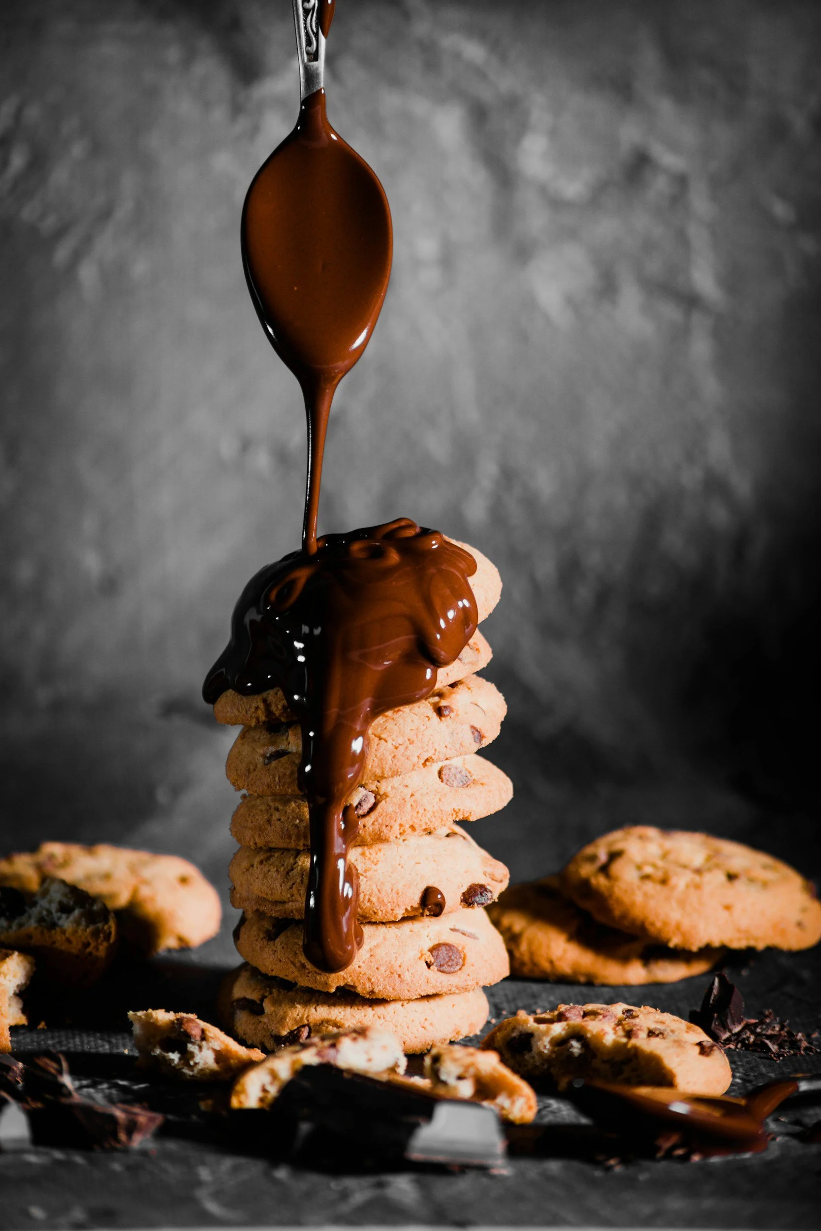 A stack of chocolate chip cookies with chocolate drizzle being poured over them, with some cookies and cookie pieces scattered around.