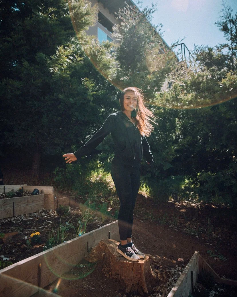 A young woman standing on a tree stump in a garden, smiling, wearing a black jacket, black pants, and sneakers, with sunlight filtering through the trees in the background.