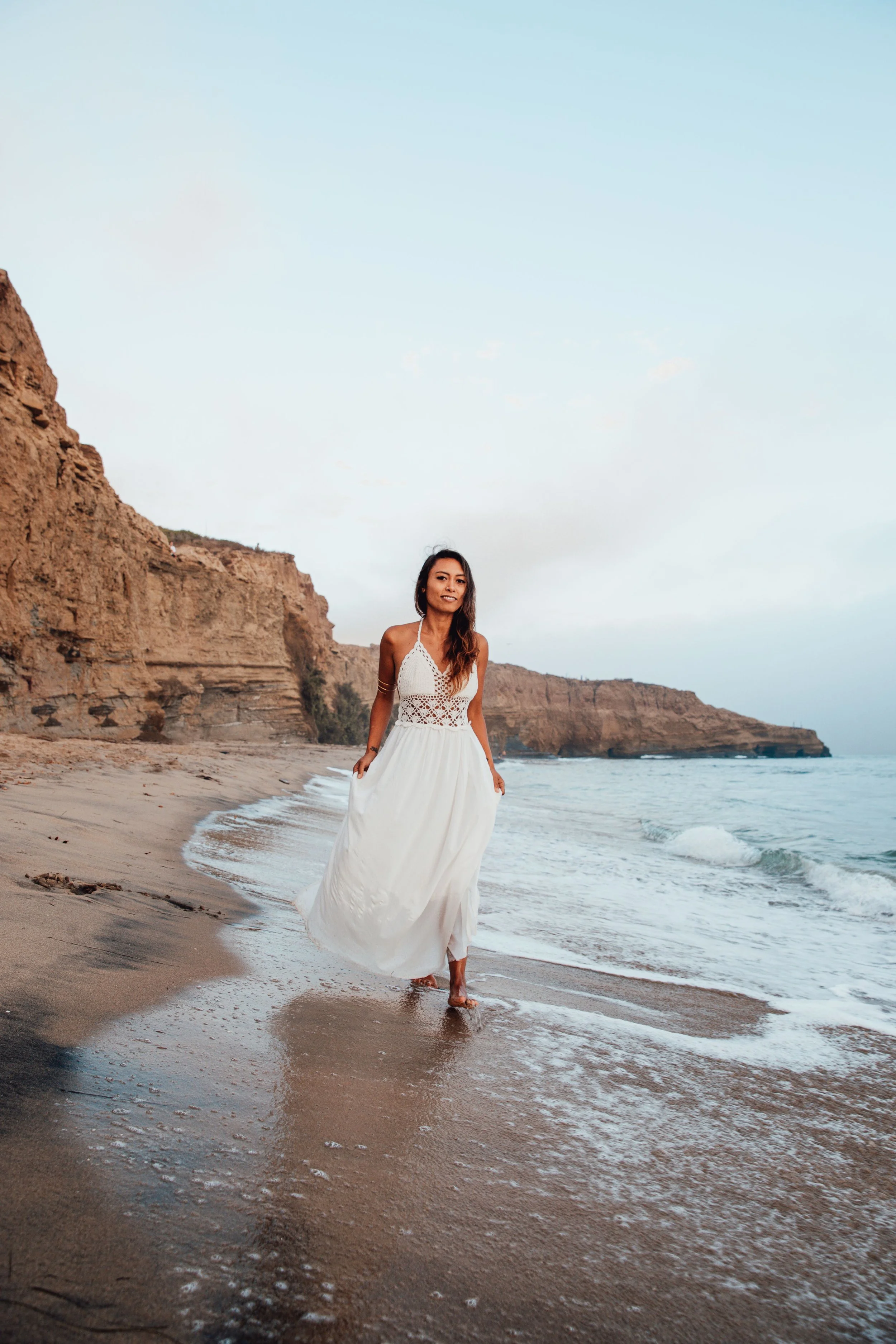Woman in a white dress walking along a sandy beach with cliffs in the background and ocean waves.