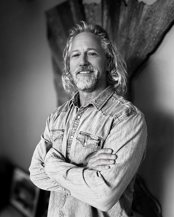 Black and white portrait of a middle-aged man with wavy hair and a beard, smiling with arms crossed, wearing a denim shirt, standing in front of a wooden canoe or boat.