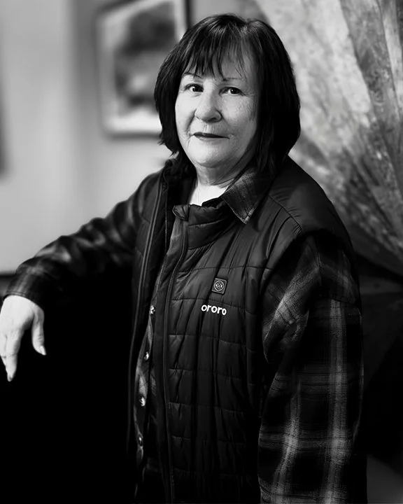 Black and white photograph of woman with short dark hair, wearing a jacket over a plaid shirt, sitting indoors with an abstract painting in the background.