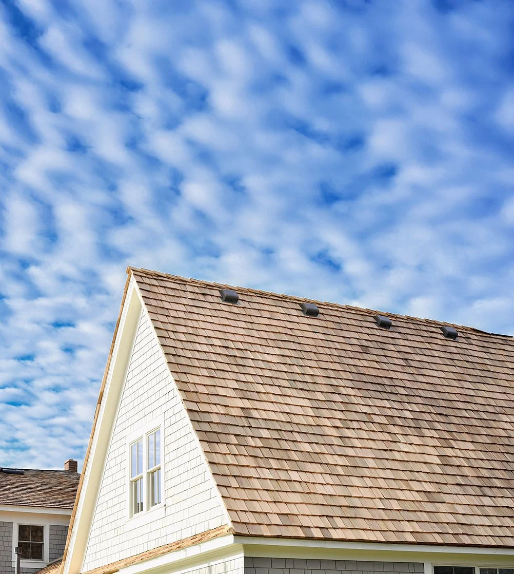 Photo of a house with a steep roof covered in brown shingles, a white wall with a window, and a partly cloudy blue sky.