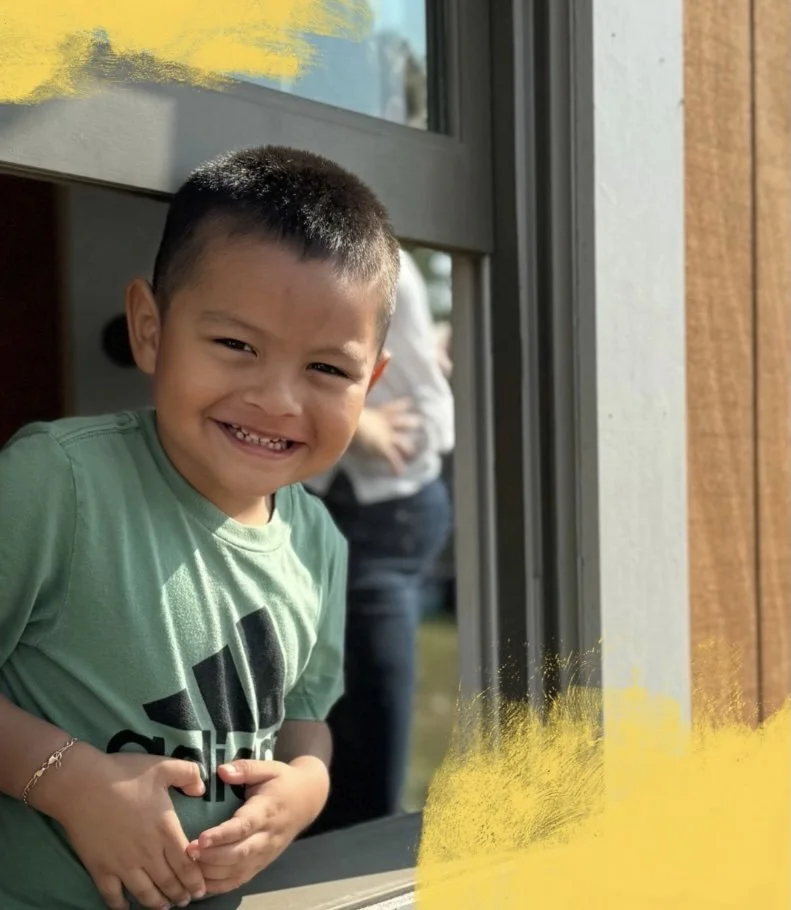 A young boy with short dark hair smiling and peeking through a window. He's wearing a light green shirt and has a bracelet on his left wrist.
