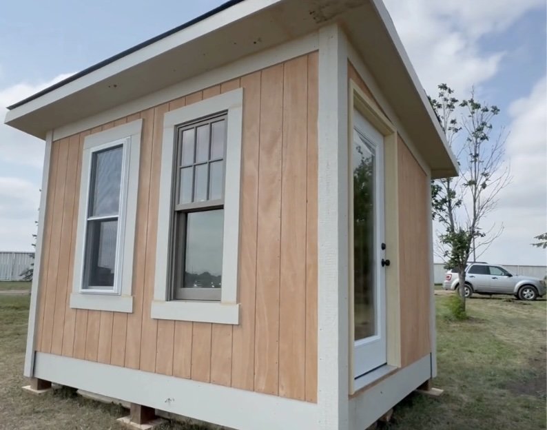Front view of small wooden house with two windows and a door, outdoor setting with grass, trees, and parked cars in background.