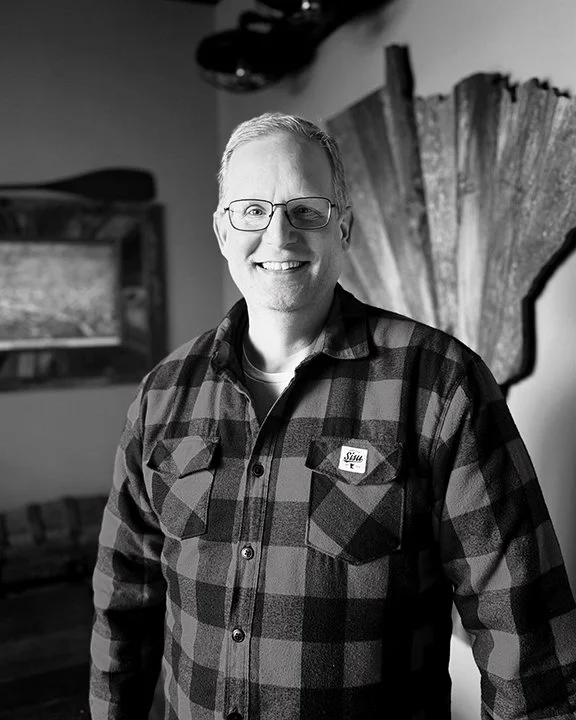 A smiling man wearing glasses and a checkered shirt in a room with art and decorative items.
