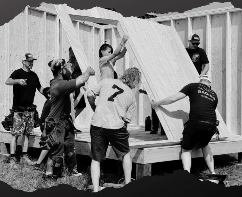 Group of people building a wooden structure, working together to set large wooden panels in place amidst a partly constructed frame.