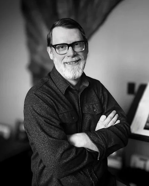 A smiling man with glasses, in a dark shirt and jacket, crossing his arms, standing in an indoor setting.