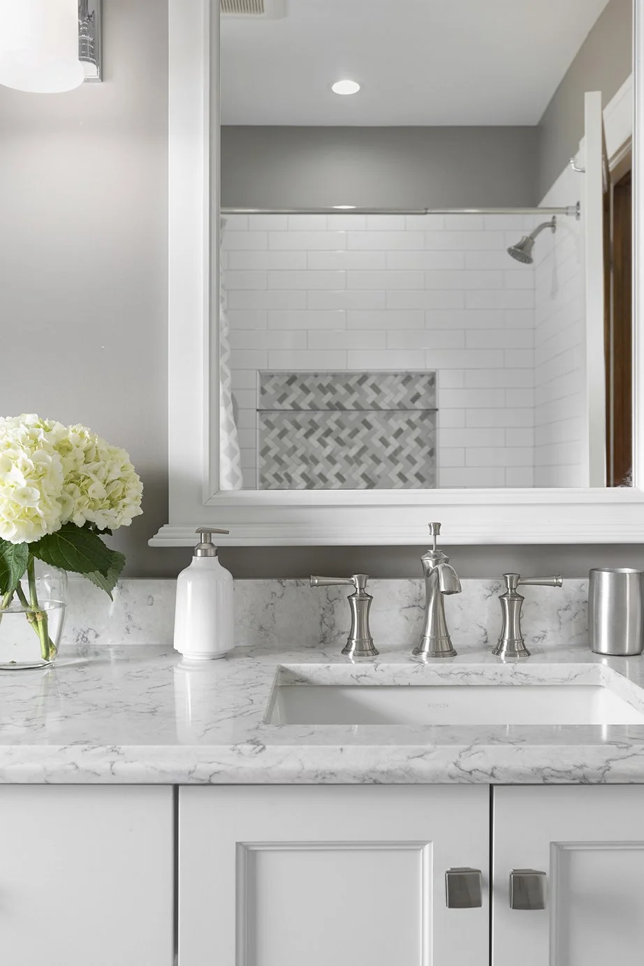 A bathroom vanity with a white marble countertop, a white sink, and fixtures, with a large mirror reflecting a shower area with white subway tiles and a decorative tiled niche.