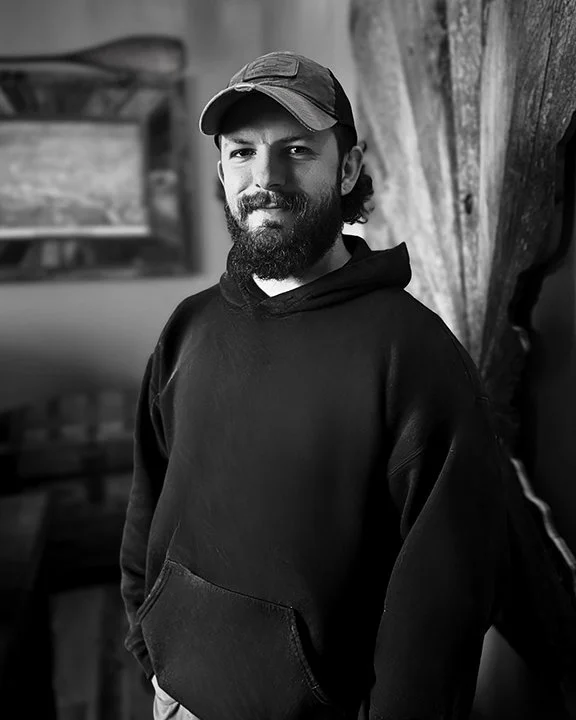 A man with a beard and mustache wearing a baseball cap and a hoodie, standing indoors next to a wooden wall.