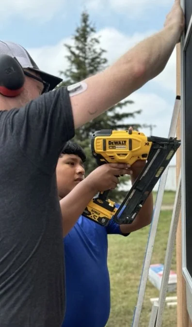 Two men working outdoors: one with a tool belt and a headset, and the other in a blue shirt holding a yellow DeWalt cordless drill on a ladder, installing or repairing something on a wall.