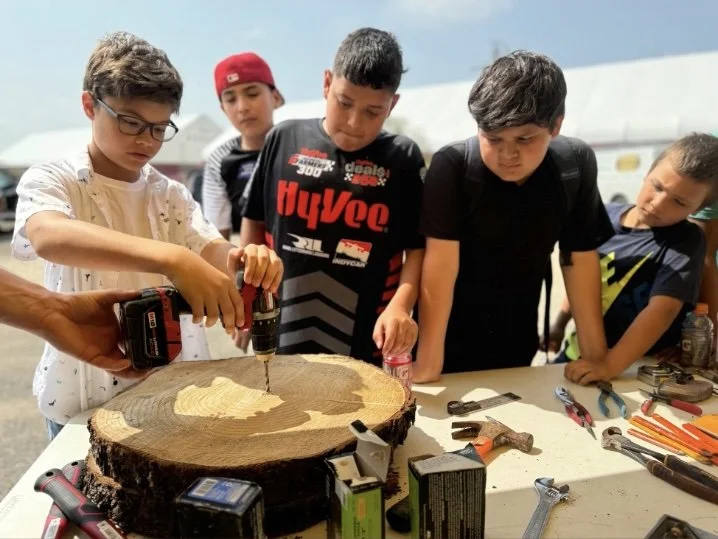 A group of boys gathered around a table outdoors, watching a boy using a drill to craft or drill into a large tree slice with a wood grain pattern, surrounded by tools.