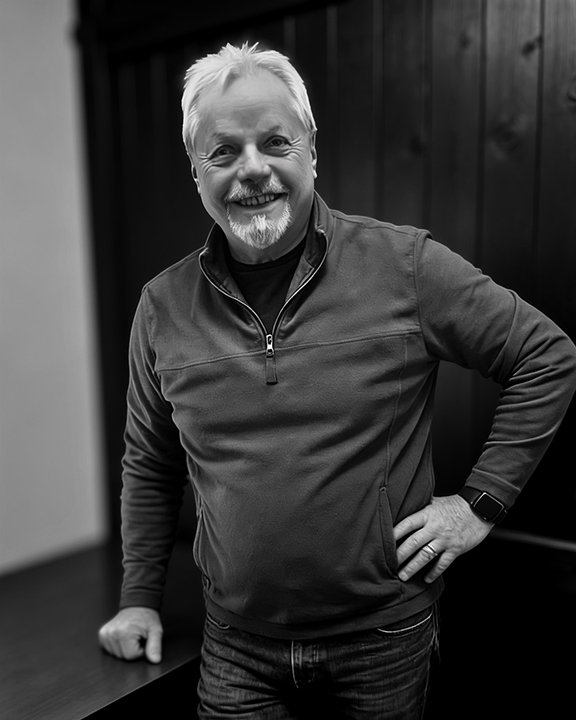 A middle-aged man with white hair and a beard smiling, wearing a zip-up jacket and a smartwatch, standing indoors against a dark wooden background.