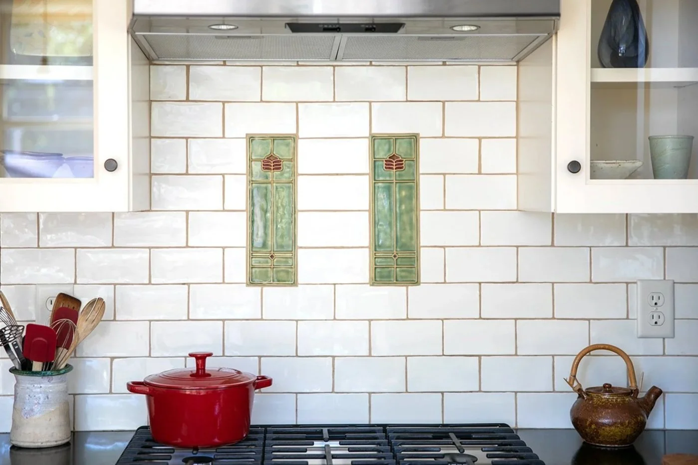 Kitchen backsplash with two decorative green and brown tiles, stove with red pot, and kitchen utensils in a container.