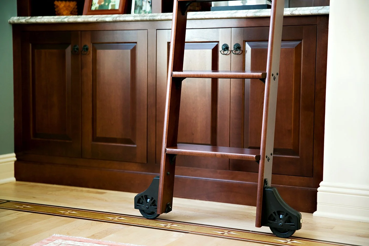 A wooden step ladder with black wheels positioned in a kitchen in front of brown cabinets, on a light-colored hardwood floor.