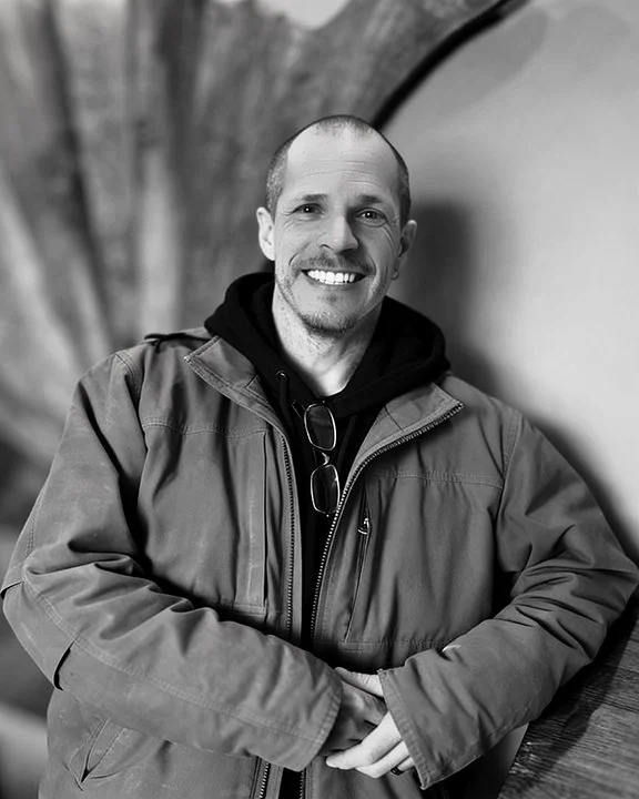 A smiling man in a jacket and hoodie standing outdoors in front of a curved wall structure.