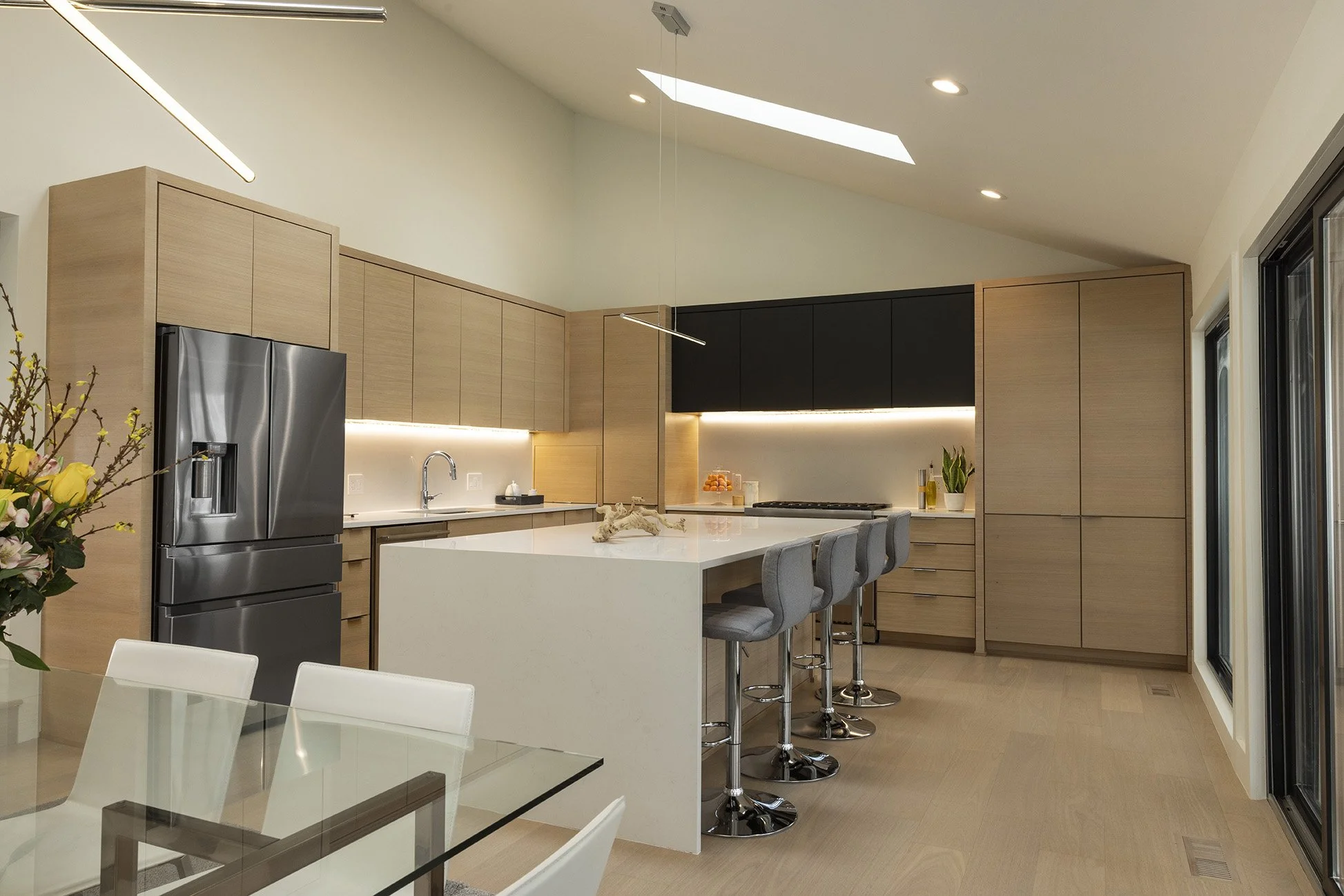 Modern kitchen with light wood cabinets, a white island, gray bar stools, stainless steel refrigerator, and large windows.