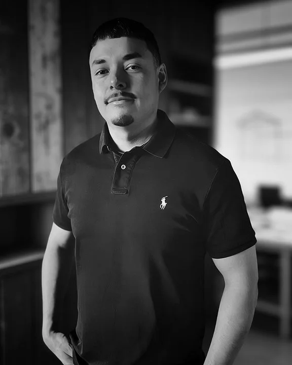 Black and white photo of a young man standing indoors, wearing a polo shirt with a small logo on the chest. He has short hair and a goatee, looking slightly to the side with a neutral expression.