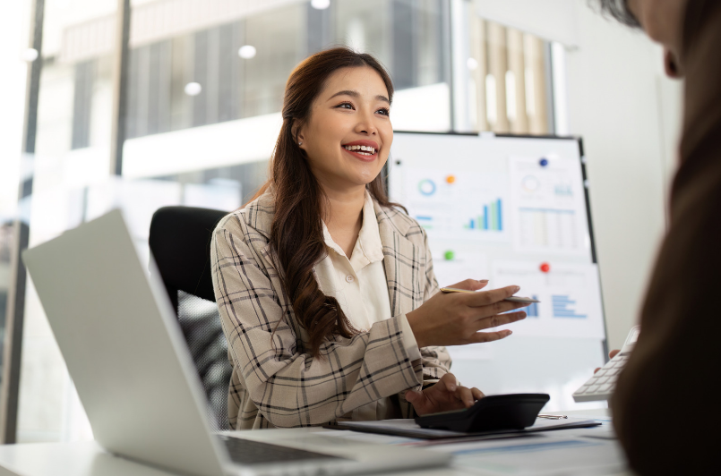 Businesswoman smiling during a meeting, holding a smartphone, with charts displayed on a screen behind her.