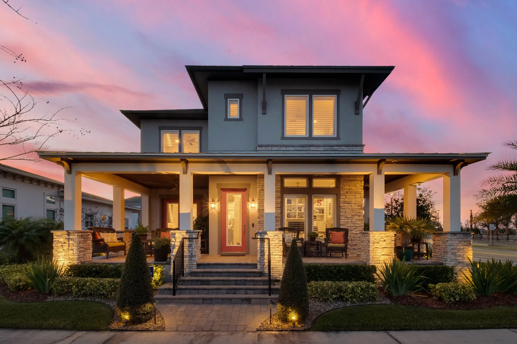 A two-story house with a covered porch, stone accents, and outdoor seating, illuminated at dusk with a colorful sky in the background.