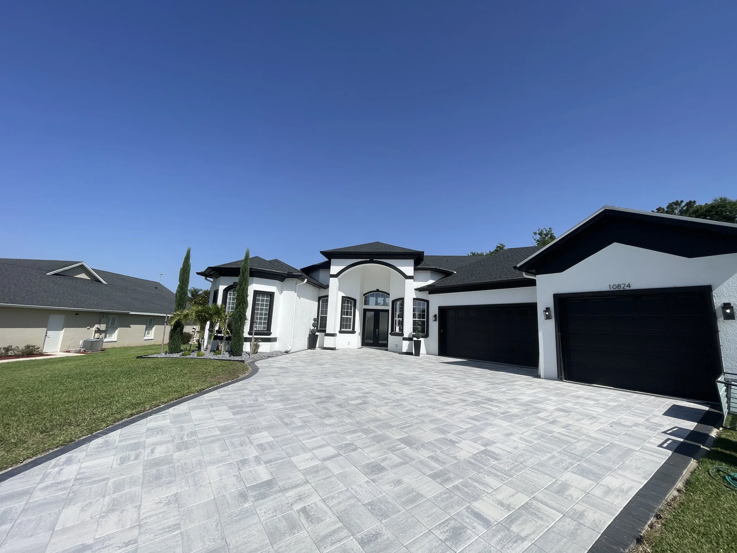Modern white house with black accents, large driveway, landscaped front yard with palm trees and cypress, black garage doors, under a clear blue sky.
