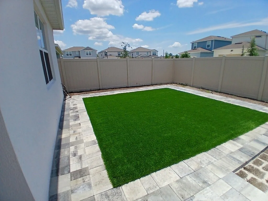 Backyard with a patch of artificial grass surrounded by stone pavers, enclosed by a beige privacy fence, under a partly cloudy blue sky.