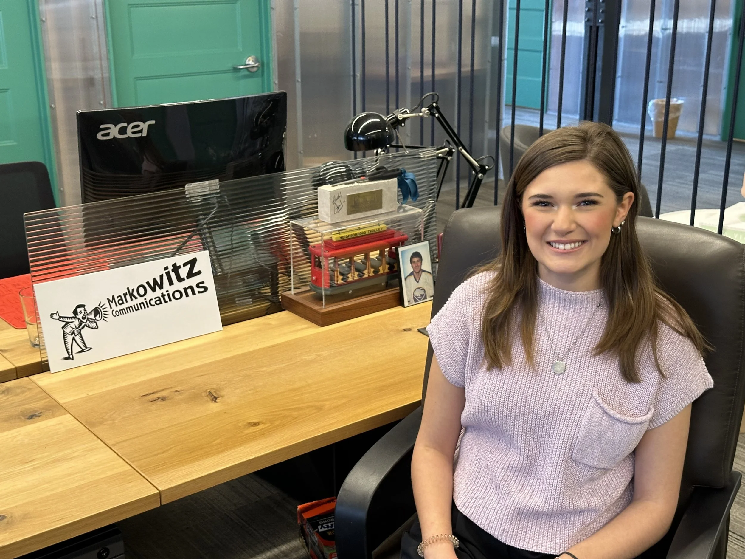 A smiling young woman with brown hair, wearing a pink short-sleeved top, sitting at a desk in an office environment. On the desk, there is a sign that reads 'Markowitz Communications', a framed photograph, and office supplies. Behind her are glass walls and a window showing an outdoor area.