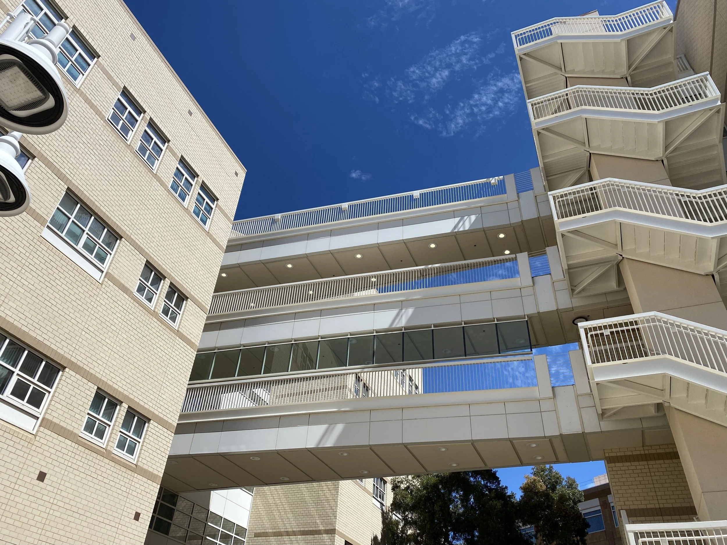 Low-angle view of a modern multi-story building with balconies, beige brick walls, and a bright blue sky with some clouds.