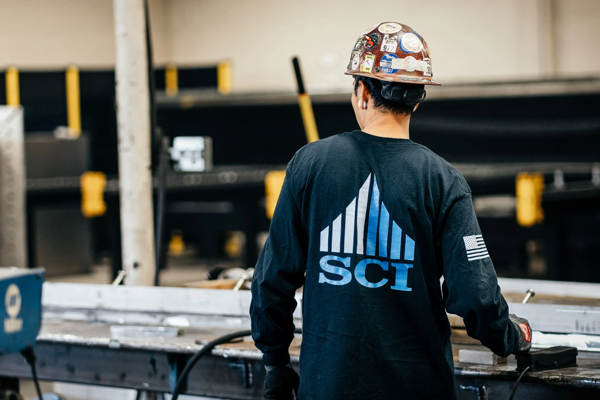 Back view of a female construction worker with short dark hair, wearing a hard hat covered in stickers and a black long sleeve shirt with a blue graphic and American flag patch, working at a construction site with metal shelving and tools.