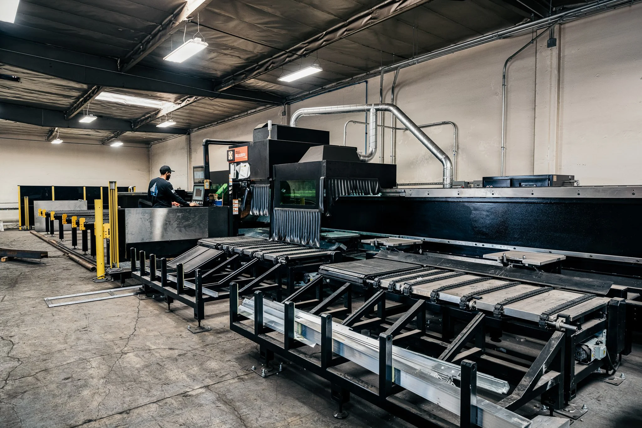 Industrial factory or warehouse with large machinery and conveyor belts. A worker wearing a cap and black shirt is operating the equipment. The room has a high ceiling with exposed pipes and pipes leading into the machine. The lighting is bright with several overhead lights.