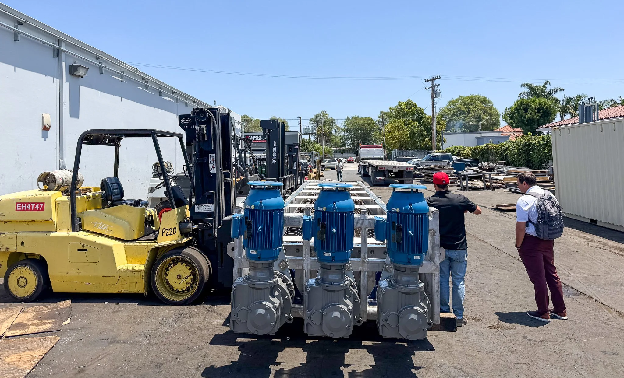 A parking lot with various equipment and vehicles, including a yellow forklift, blue water meters, trucks, and people walking around on a sunny day with trees and buildings in the background.
