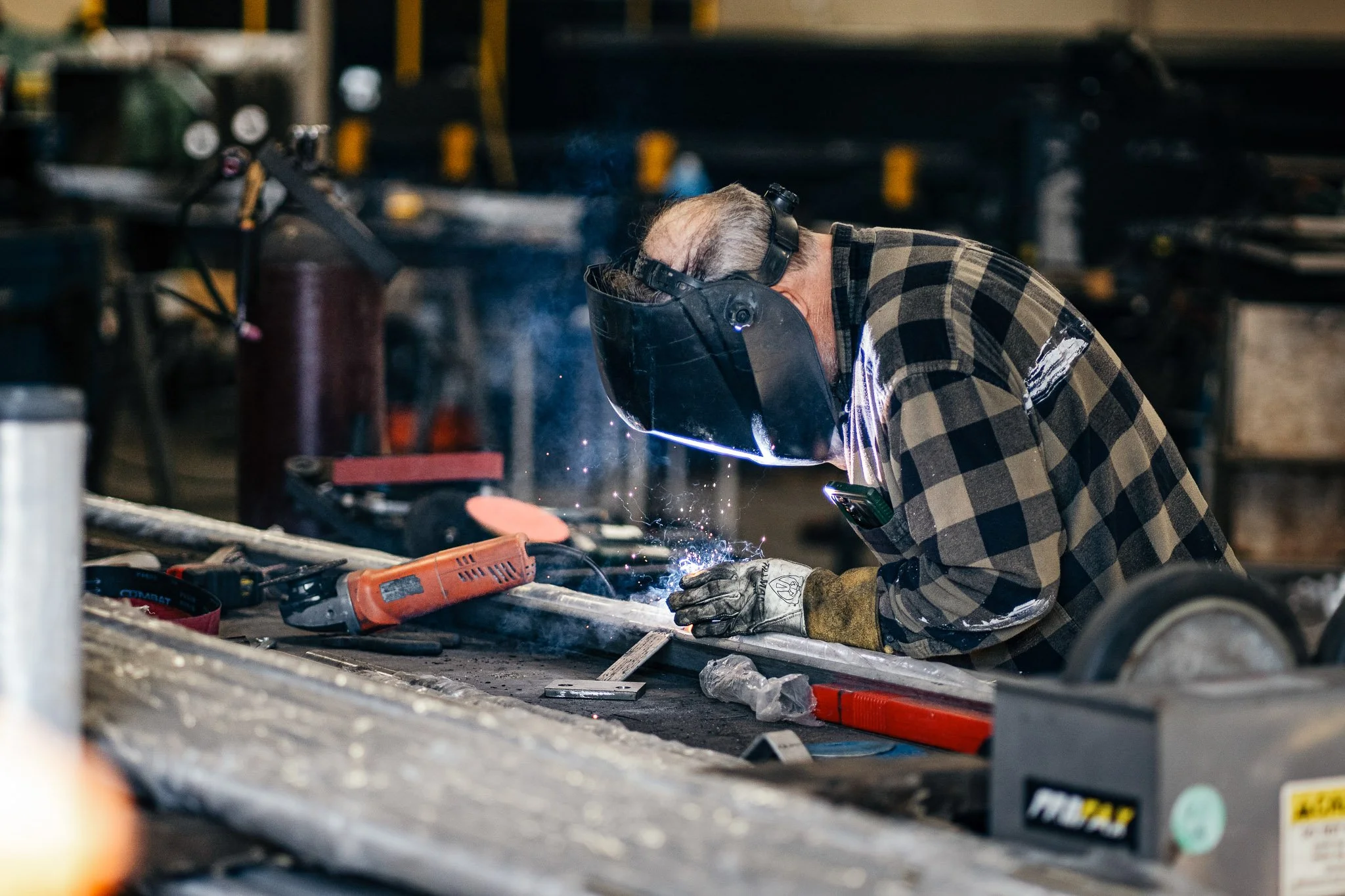 A person welding at a workbench in a workshop, wearing safety glasses and ear protection, surrounded by various tools and equipment.