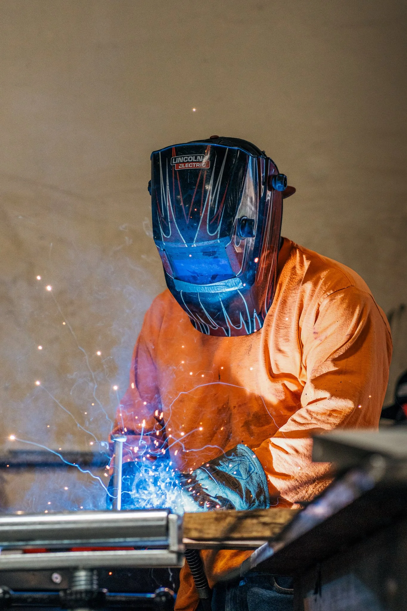 A person wearing an orange shirt and a welding mask is welding metal, creating bright sparks and blue light.